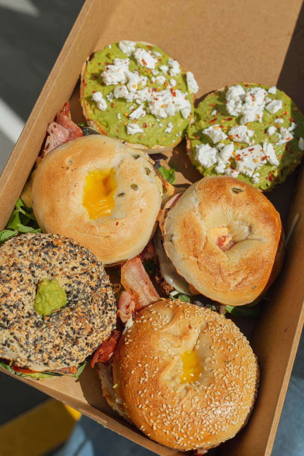 A Dirty Deli box of filled bagels and open avocado bagels with feta